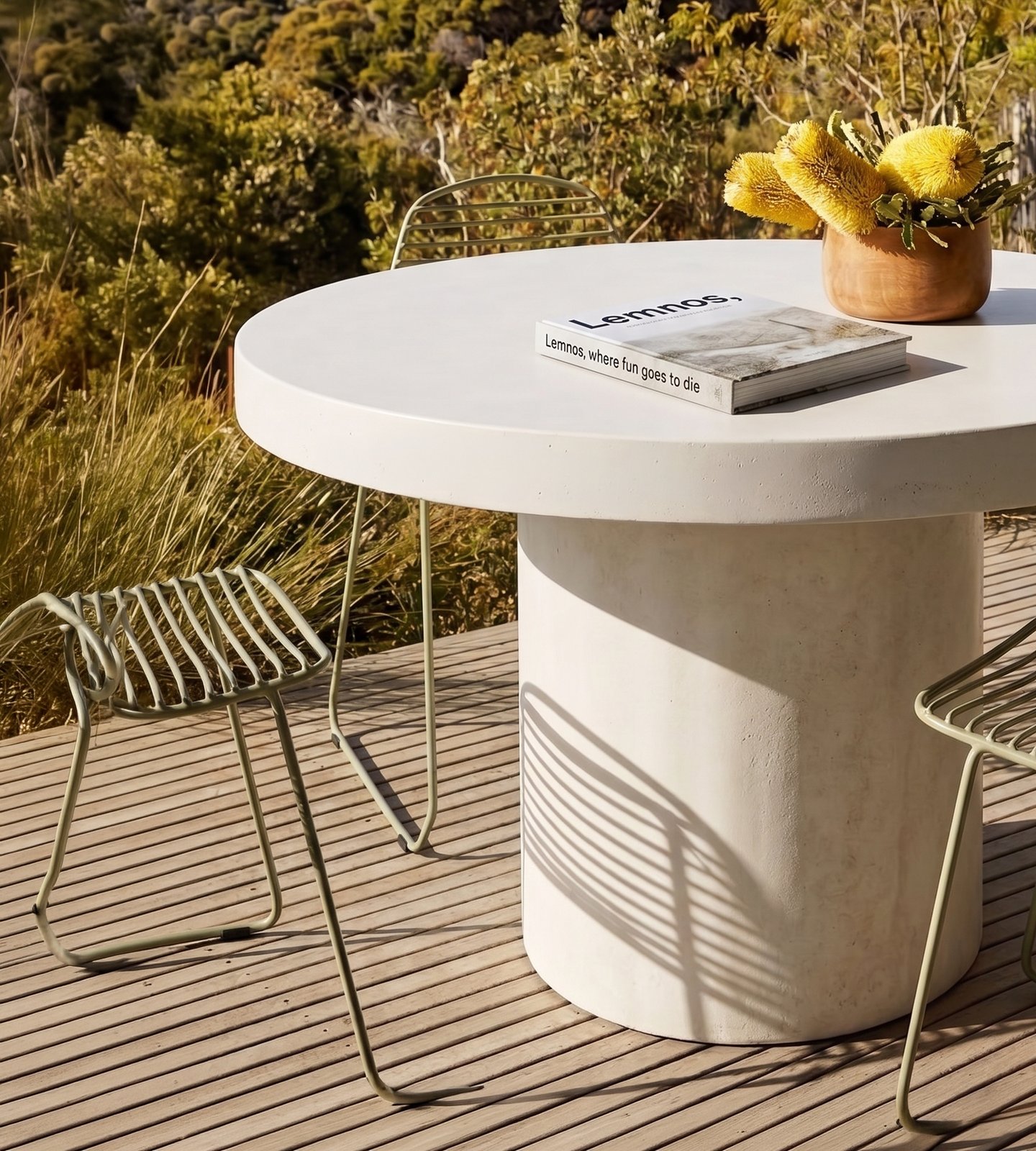 Athina round concrete dining table on a coastal deck with green wire chairs, yellow banksia floral arrangement and bush view