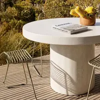 Athina round concrete dining table on a coastal deck with green wire chairs, yellow banksia floral arrangement and bush view