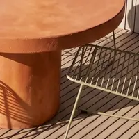 Terracotta-pigmented Athina round concrete dining table base and edge detail on a timber deck with olive wire chair and palm-frond shadow