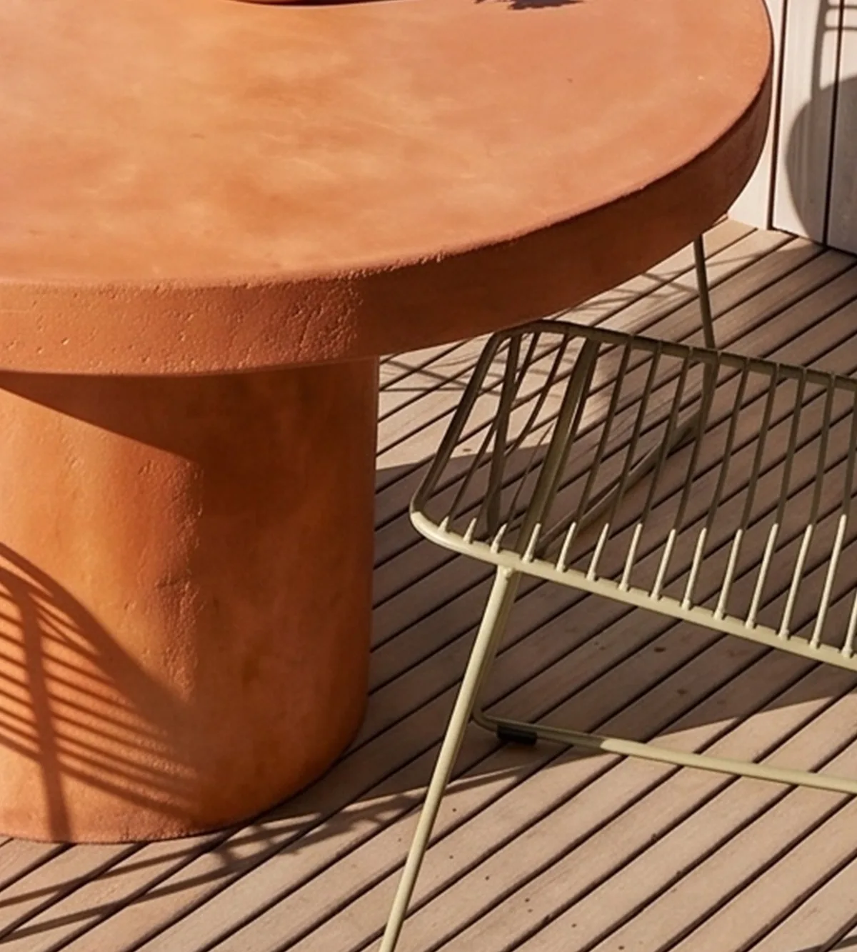 Terracotta-pigmented Athina round concrete dining table base and edge detail on a timber deck with olive wire chair and palm-frond shadow