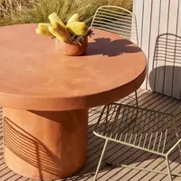 Terracotta-pigmented Athina round concrete dining table on a coastal deck with olive wire chairs, yellow banksia arrangement and dune-grass view