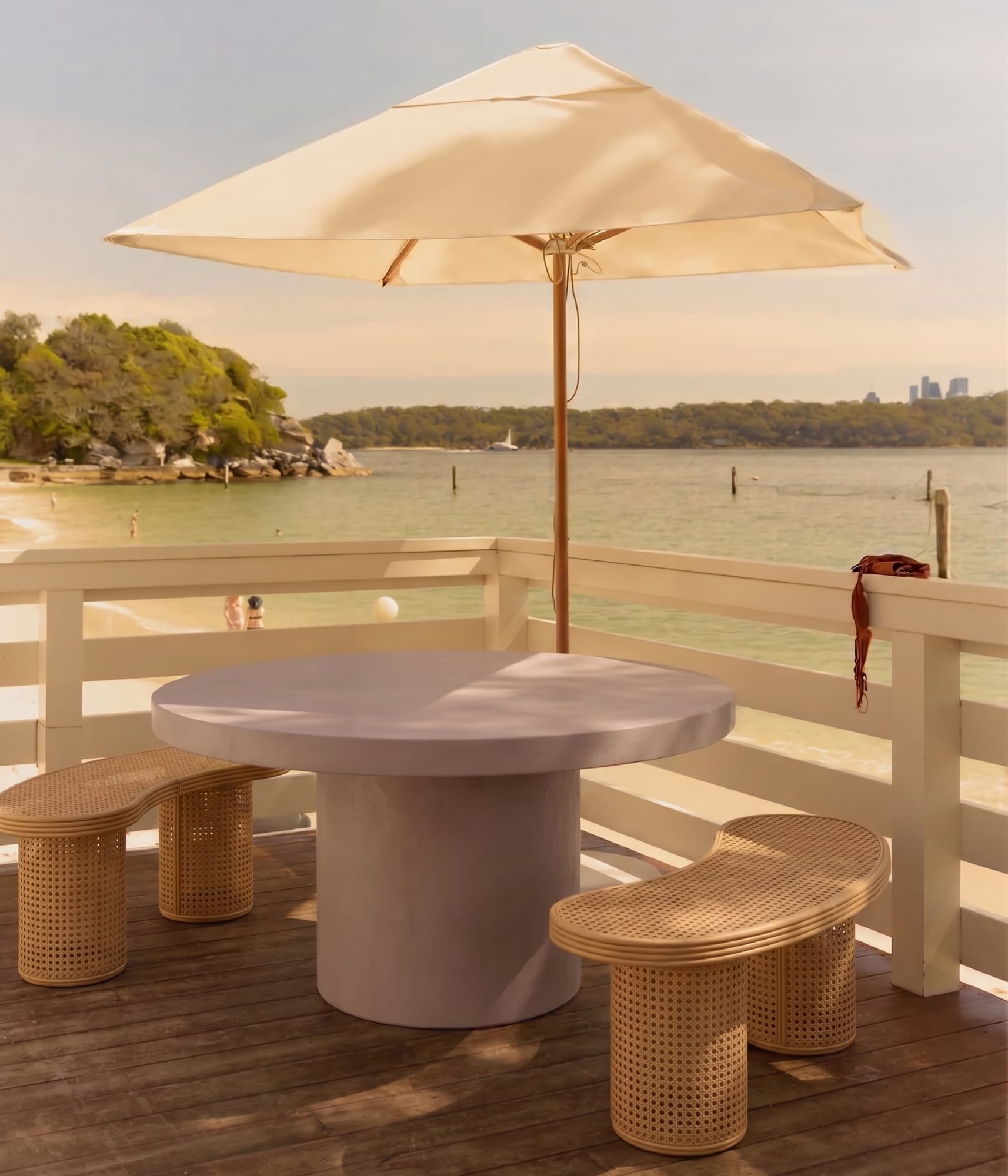 Athina round bone-white concrete dining table on a beachside deck under a cream umbrella, paired with rattan crescent benches, harbour view beyond