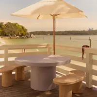 Athina round bone-white concrete dining table on a beachside deck under a cream umbrella, paired with rattan crescent benches, harbour view beyond