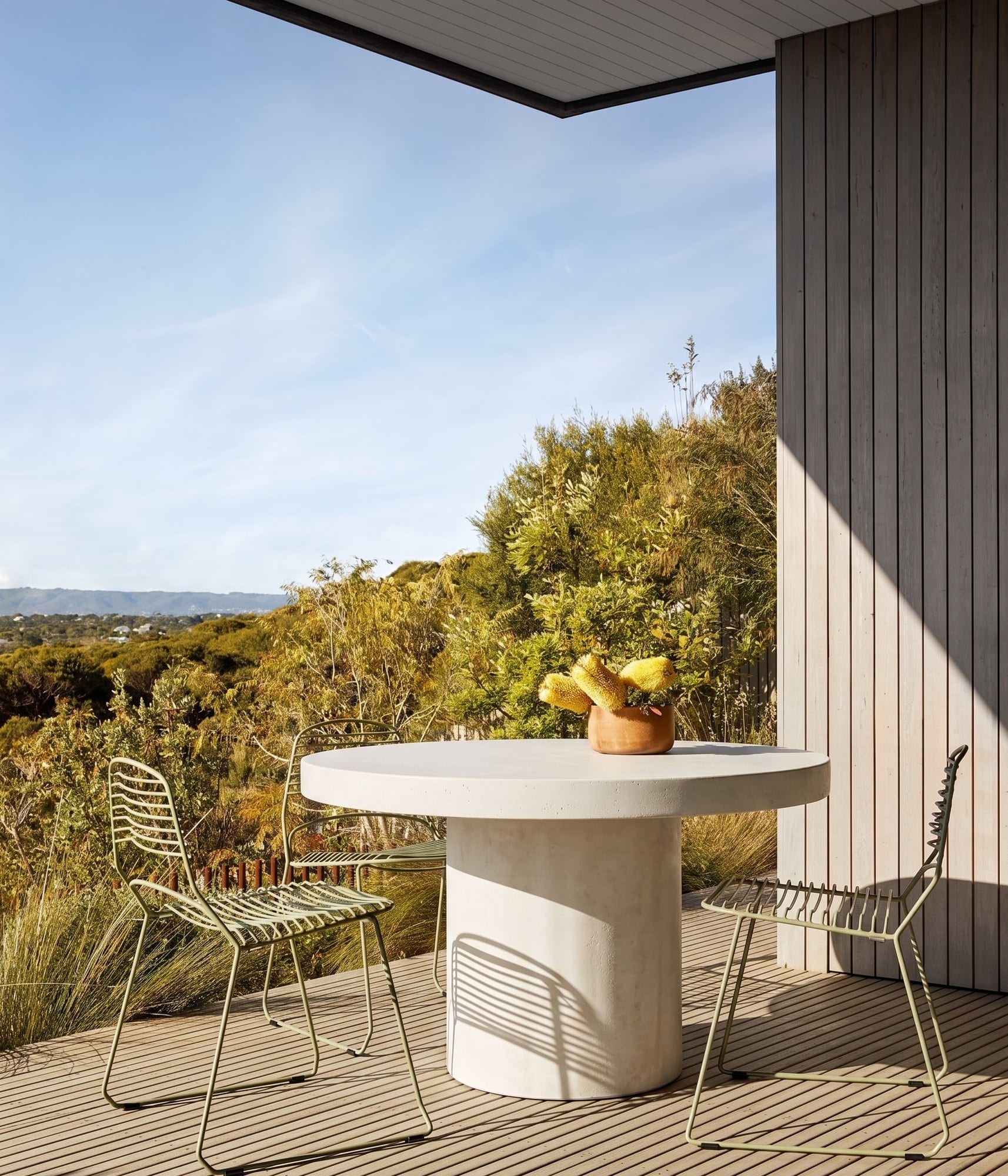 Athina round bone-white concrete dining table on a Mornington Peninsula timber deck with sage-green wire chairs and a banksia arrangement, coastal bushland beyond
