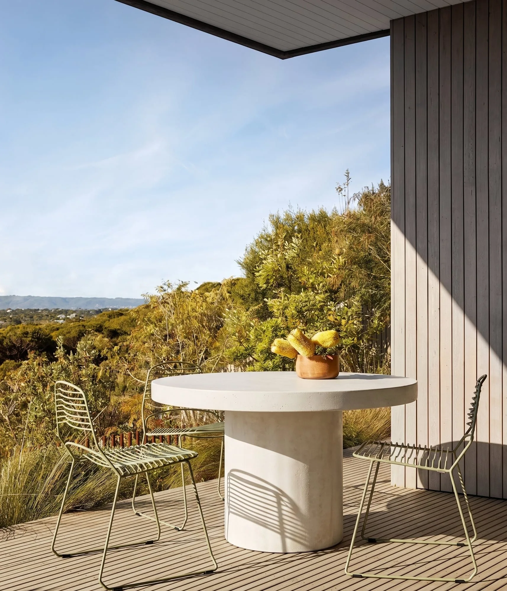 Athina round bone-white concrete dining table on a Mornington Peninsula timber deck with sage-green wire chairs and a banksia arrangement, coastal bushland beyond