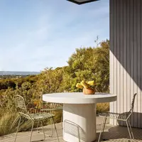 Athina round bone-white concrete dining table on a Mornington Peninsula timber deck with sage-green wire chairs and a banksia arrangement, coastal bushland beyond