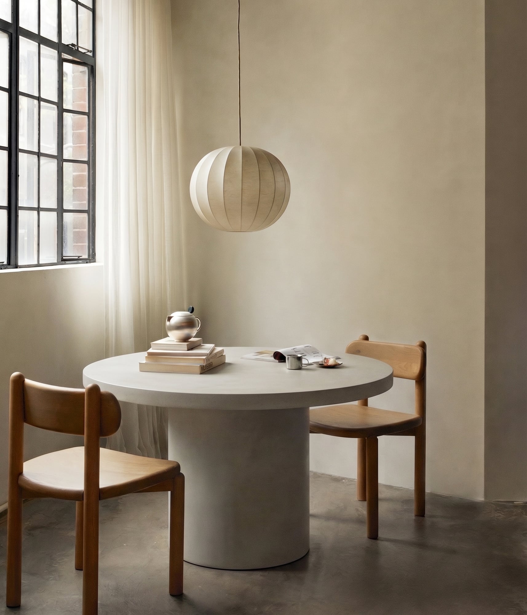 Athina round bone-white concrete dining table in a sun-washed interior with a paper sphere pendant, sheer curtains, two timber chairs and a stack of art books
