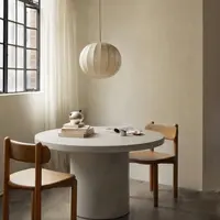 Athina round bone-white concrete dining table in a sun-washed interior with a paper sphere pendant, sheer curtains, two timber chairs and a stack of art books