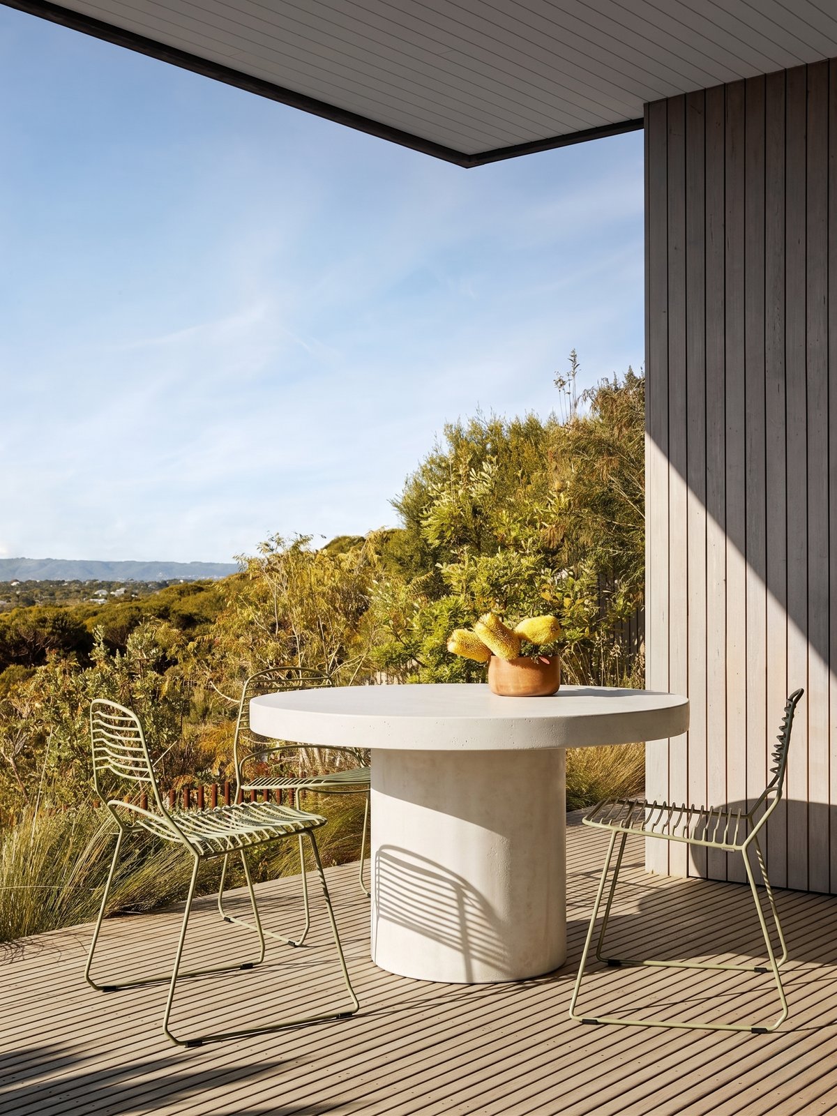 Athina round concrete dining table on a covered deck with ocean and bushland views, green wire chairs and yellow banksia arrangement