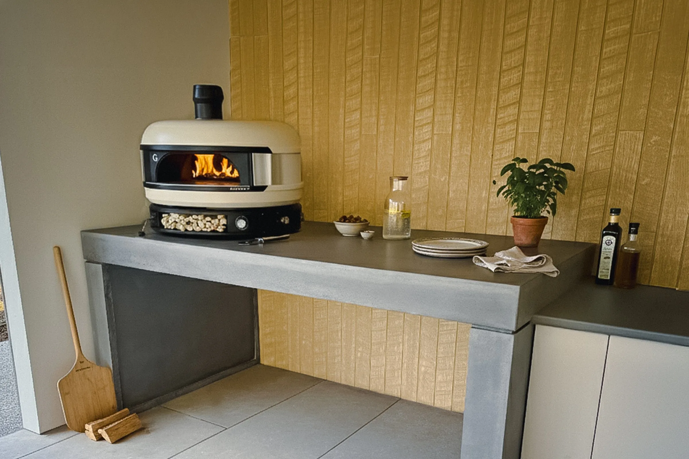 A SnapCo concrete outdoor kitchen benchtop with a Gozney wood-fired pizza oven landing, oak-batten splashback, terracotta basil pot, olive oil, and a pizza peel