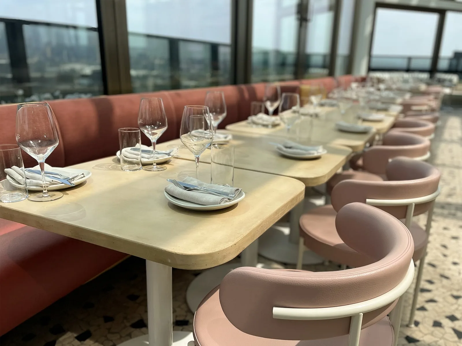 A long bone white concrete restaurant table run along a peach velvet banquette at The Beverly Rooftop in South Yarra, set for service with glassware, linen and silverware, terrazzo floor underneath and the Melbourne skyline through the window wall