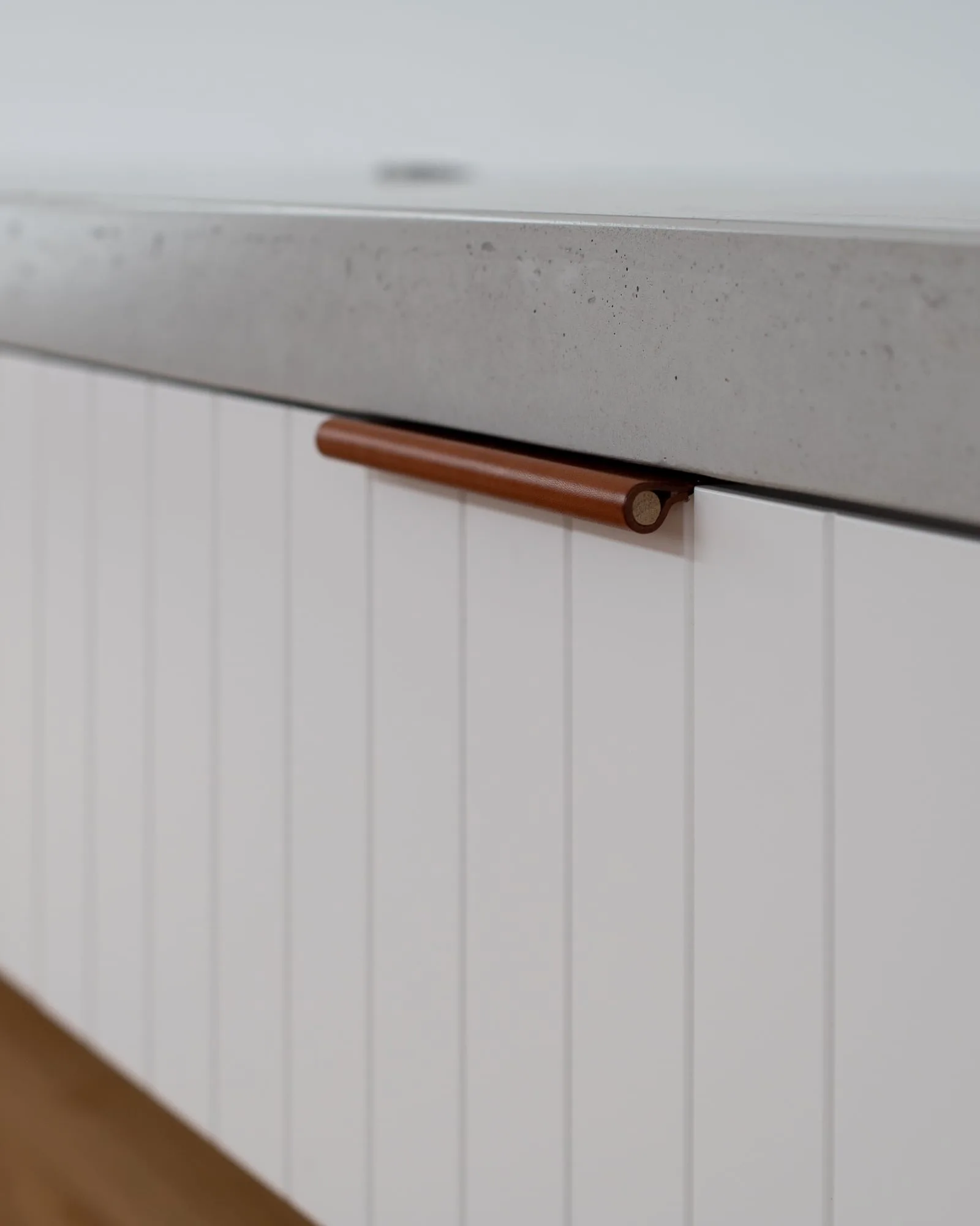 Close-up of a grey concrete benchtop edge meeting white v-groove cabinetry with leather tab pulls