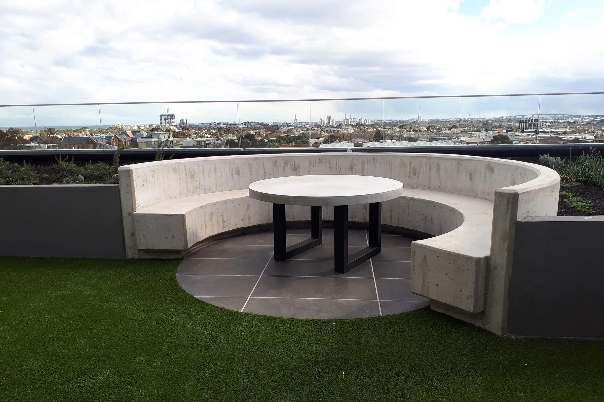 A SnapCo commercial concrete seating installation: curved cast-concrete banquette wrapping a round concrete table on a Melbourne rooftop terrace, glass balustrade, city skyline beyond