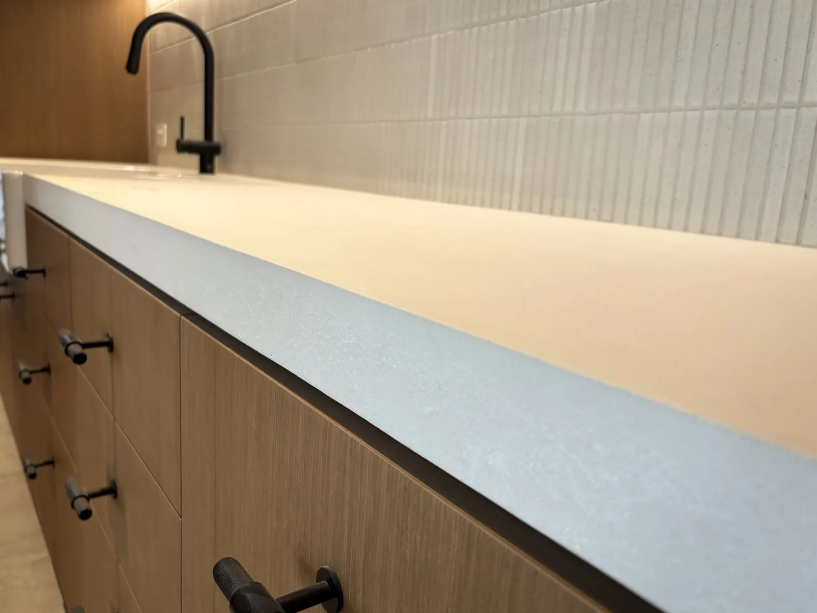 A SnapCo custom-built bone white concrete kitchen benchtop edge against light oak drawer fronts with a black tap and ribbed splashback tile