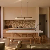 Farmhouse white concrete kitchen island with oak A-frame base, beneath a brass linear pendant against a warm checker tile splashback, dining bench and stools