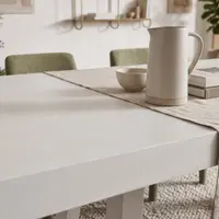 White IronCross concrete dining table in a light-filled interior with brass shelving, olive velvet chairs and fiddle-leaf fig