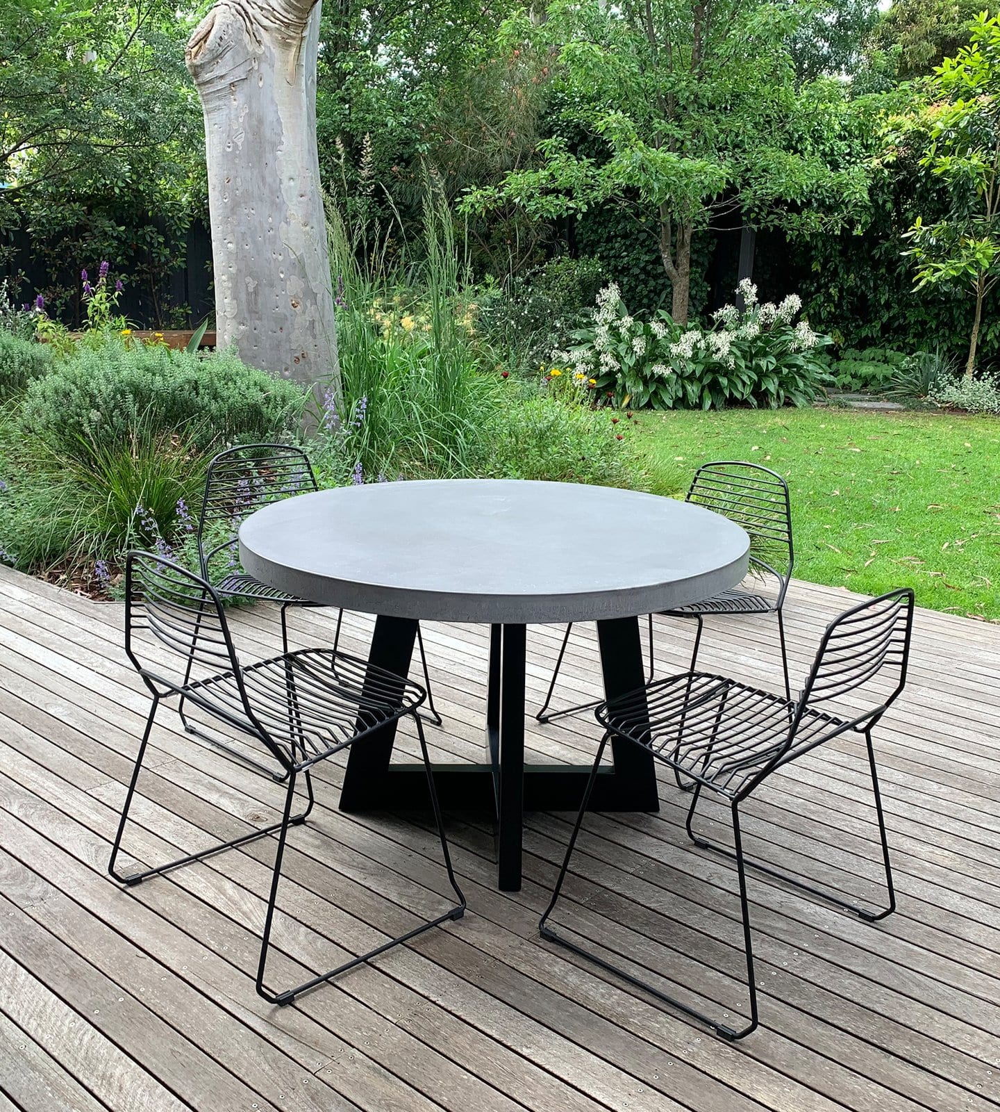 Round IronCross concrete dining table on a timber deck overlooking a lush cottage garden with black wire chairs