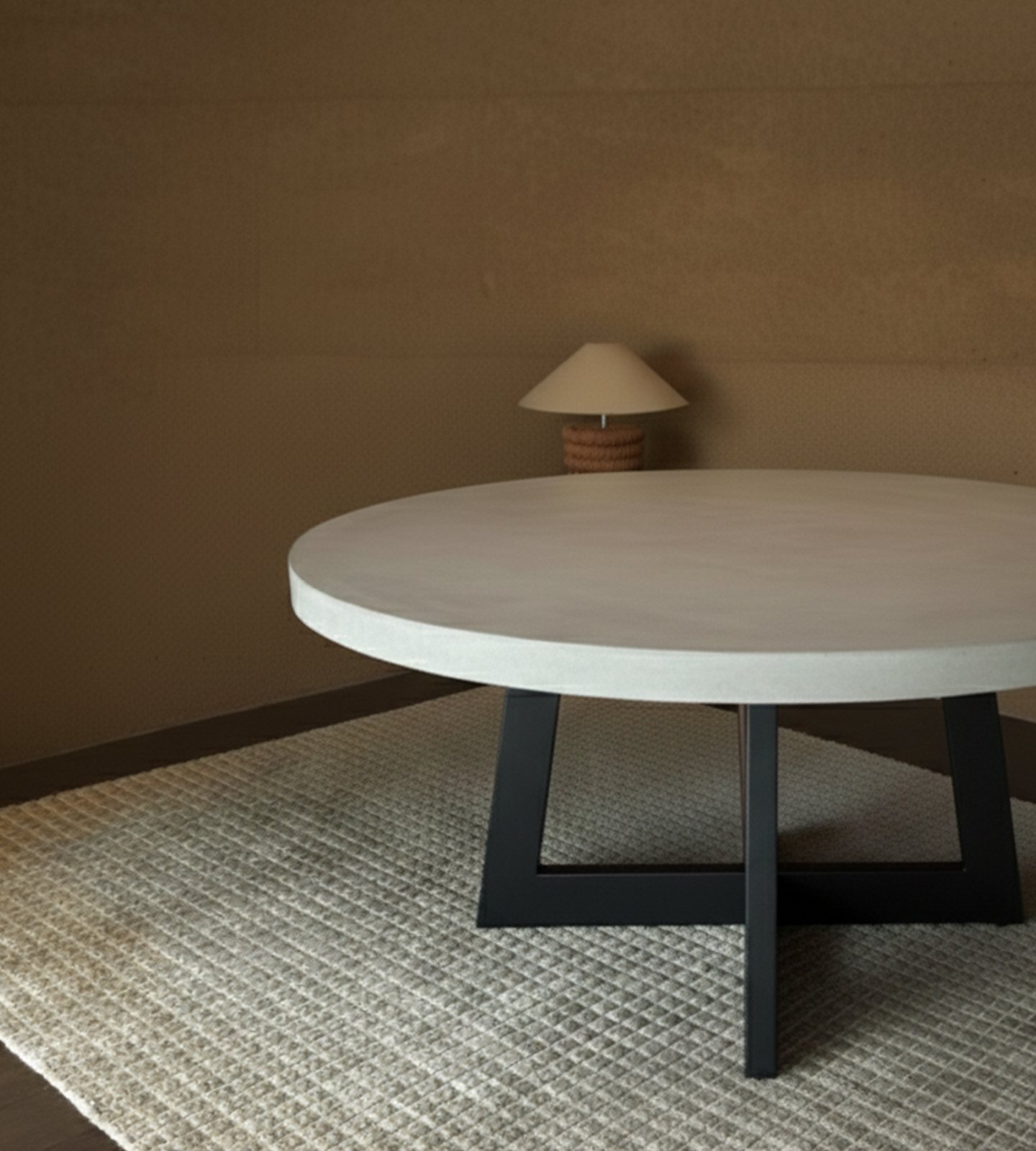 Round IronCross concrete dining table on a steel cross-base in a warm interior with paper-shade table lamp