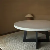 Round IronCross concrete dining table on a steel cross-base in a warm interior with paper-shade table lamp