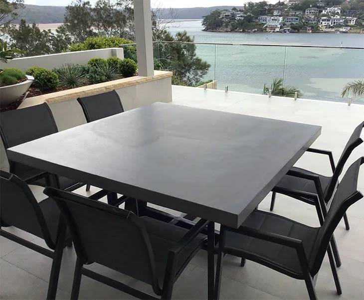 Square IronCross concrete dining table on a coastal terrace with water and waterfront homes in the background, framed by black mesh dining chairs