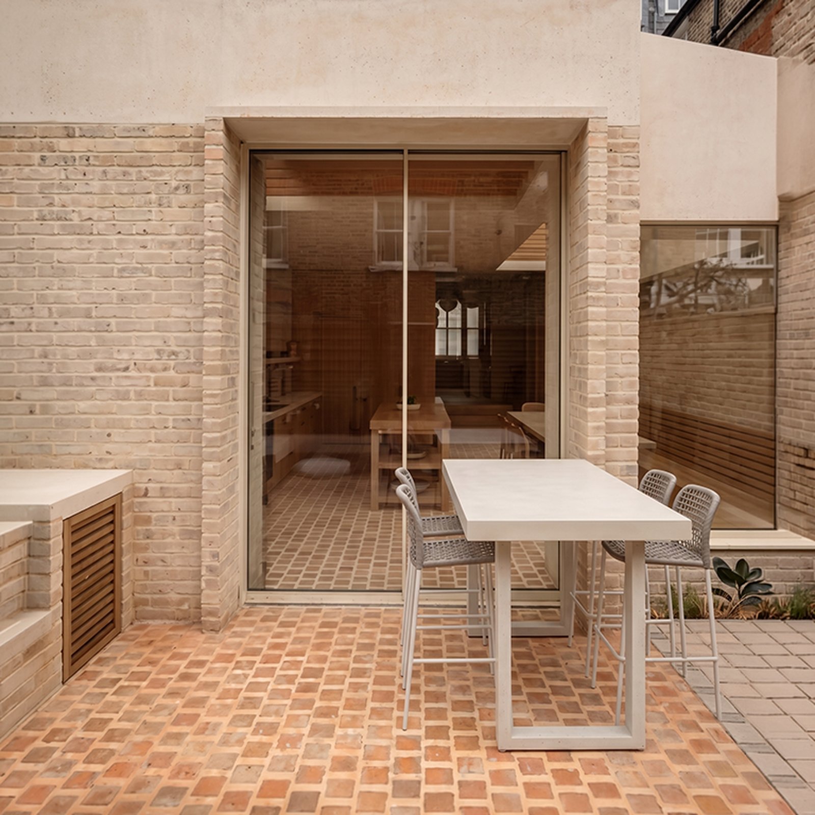 IronStone white concrete bar table on a London brick courtyard outside a tall sliding glass door, with grey rope-back bar stools