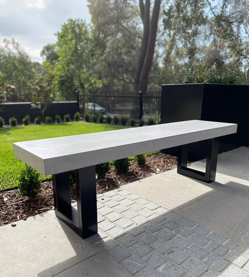 IronStone concrete bench seat with black steel sled legs on pavers, beside a manicured garden with hedge and lawn