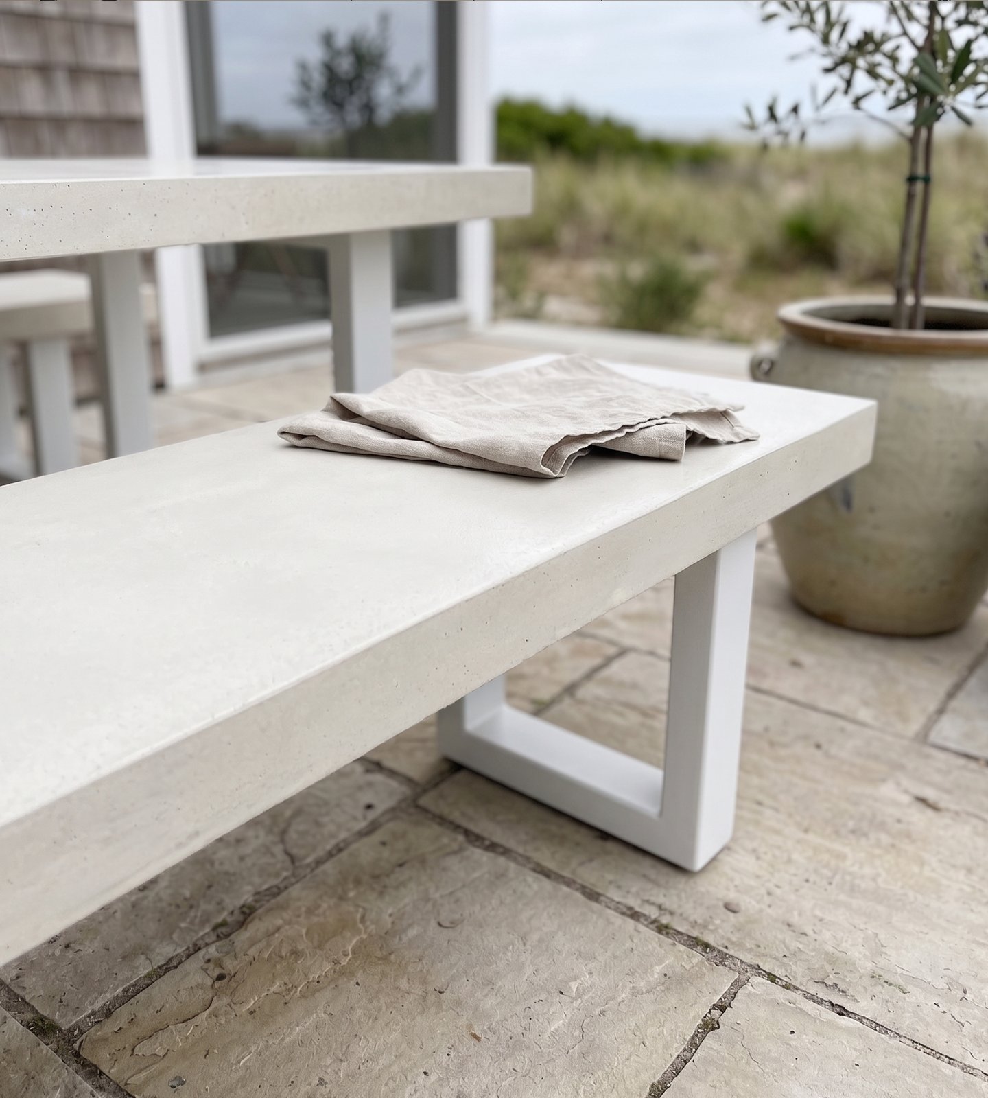 Close-up of a white IronStone concrete bench on a beach-house flagstone patio with olive tree in a stoneware pot