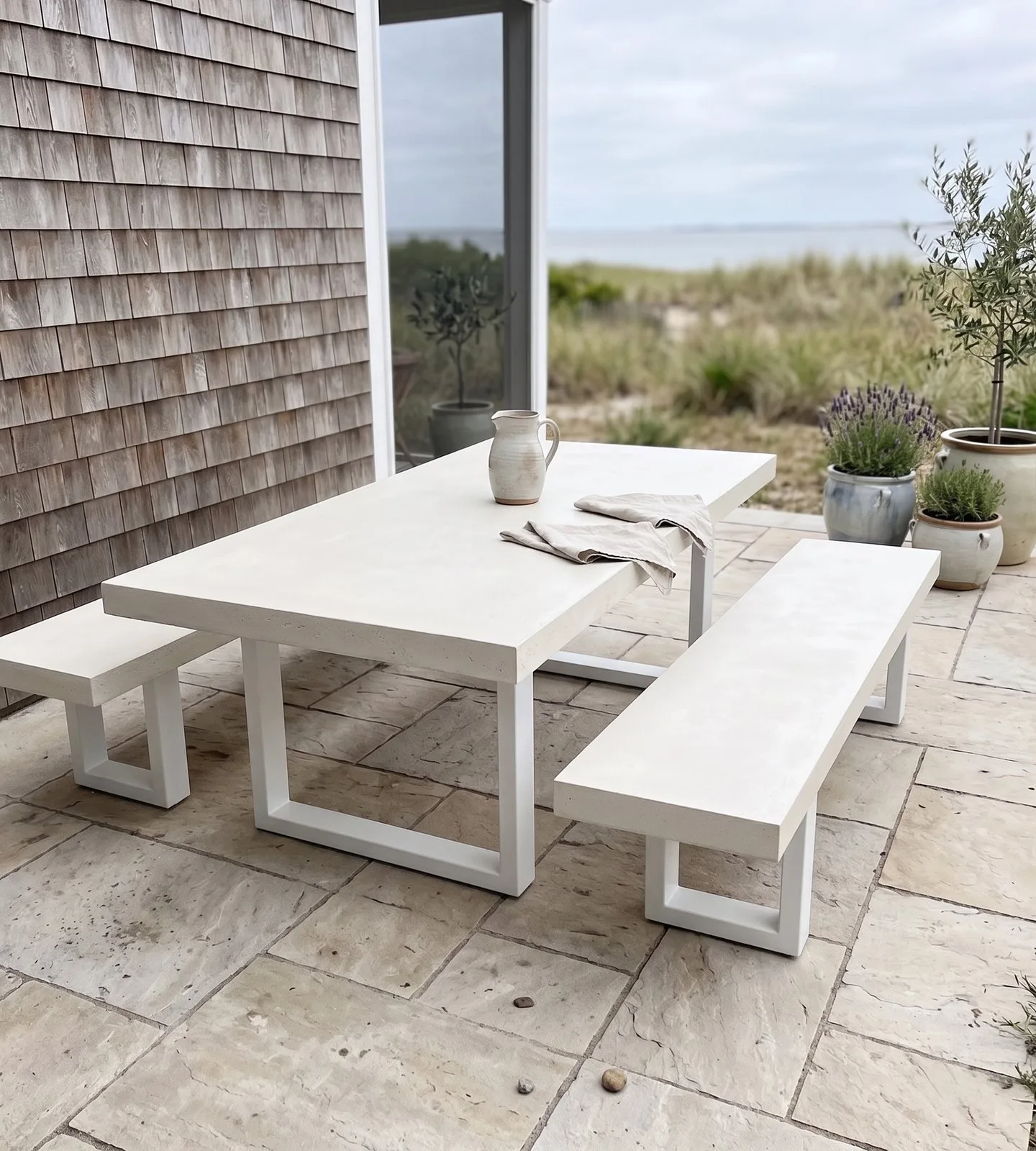 White IronStone concrete dining table and bench seat on a beach-house flagstone patio with shingle wall and olive trees