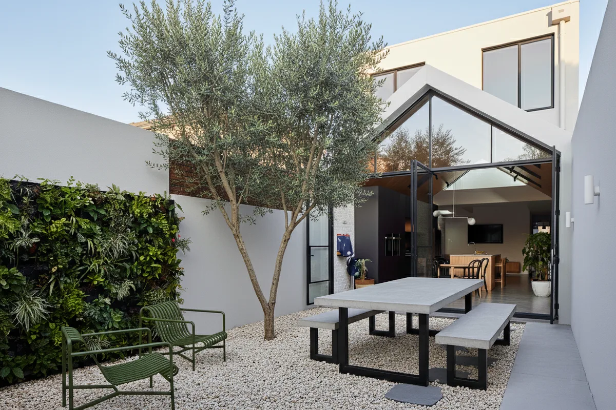 IronStone concrete dining table with matched bench seats in a courtyard with olive tree, vertical green wall and white pebbles, beside a contemporary home