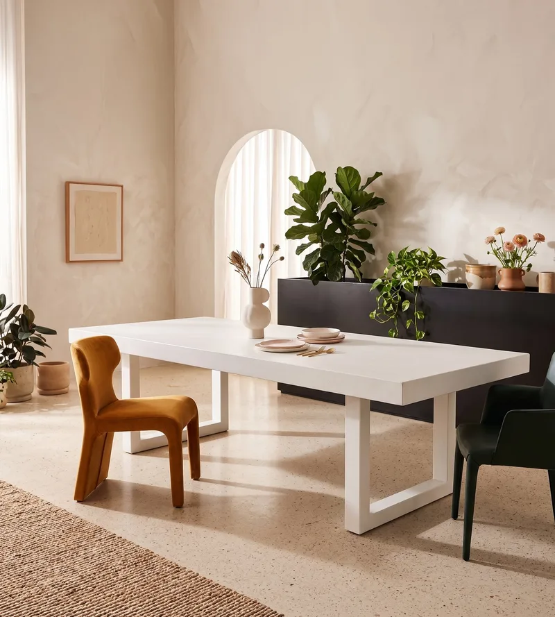 White IronStone concrete dining table in a sunlit plaster interior with arched window, fiddle-leaf fig and mustard wing chair