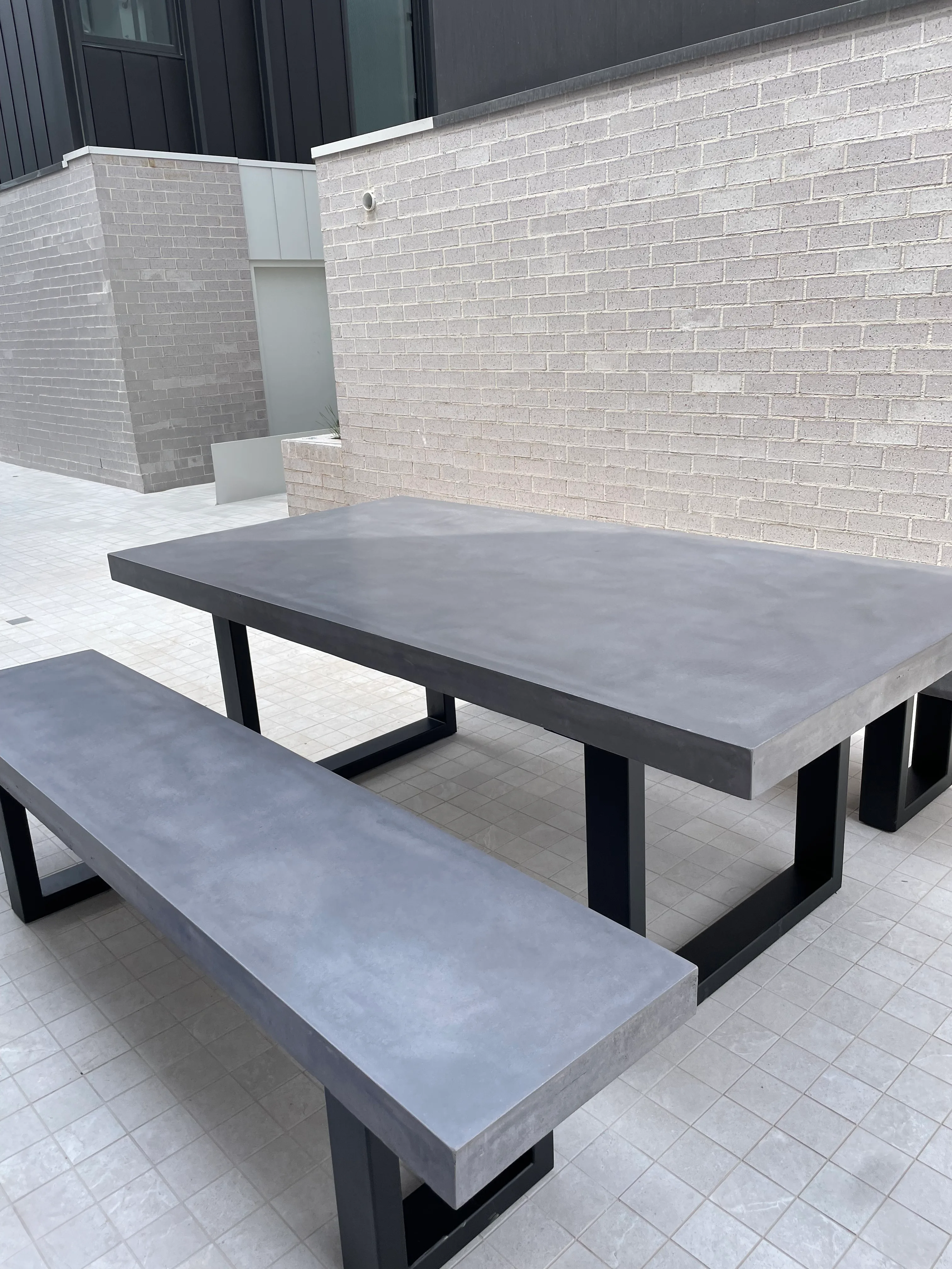 A SnapCo IronStone concrete bench seat paired with a matched concrete dining table in the communal BBQ area at 93 Earl Street Kew, set on a pale travertine paving floor with white brick perimeter walls