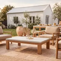 Laconian white concrete coffee table with oak base on a terracotta patio, with boucle armchairs and farmhouse beyond
