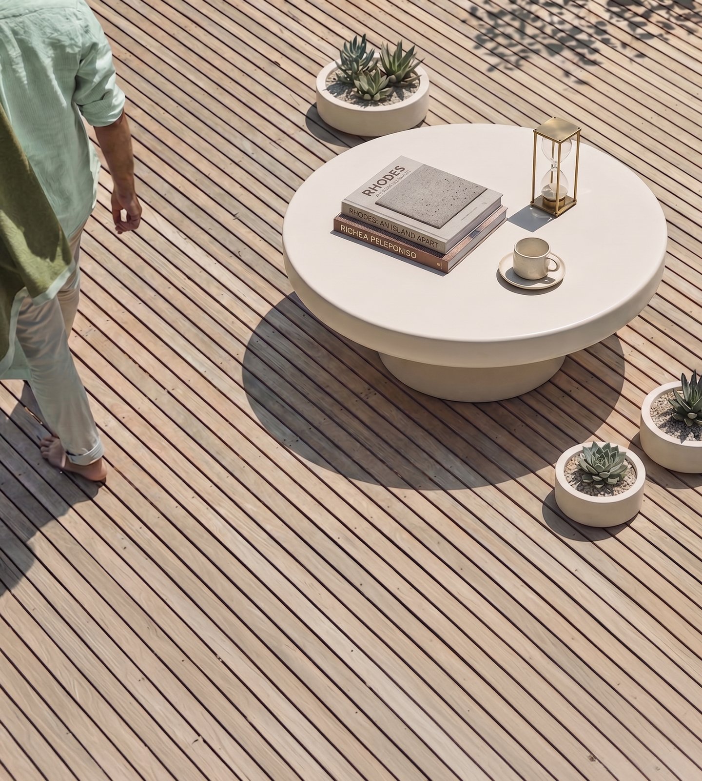 Milano white concrete coffee table on a sunlit timber deck with travel books, brass hourglass, espresso cup and trio of concrete succulent planters, overhead view with figure in linen