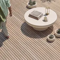 Milano white concrete coffee table on a sunlit timber deck with travel books, brass hourglass, espresso cup and trio of concrete succulent planters, overhead view with figure in linen