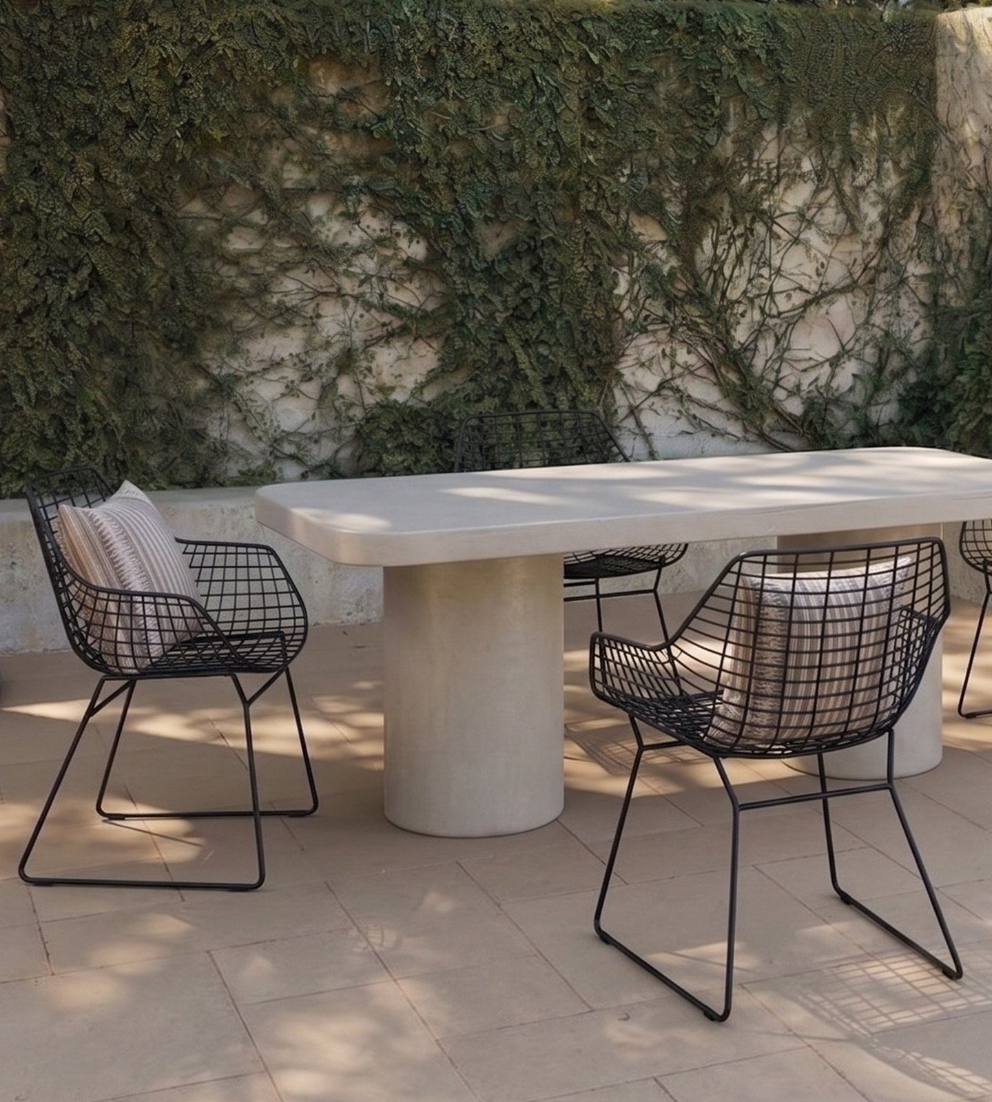 Milano rectangular white concrete dining table on twin cylindrical concrete pedestals in a sunlit Mediterranean courtyard with black wire armchairs, striped cushions and an ivy-clad plaster wall