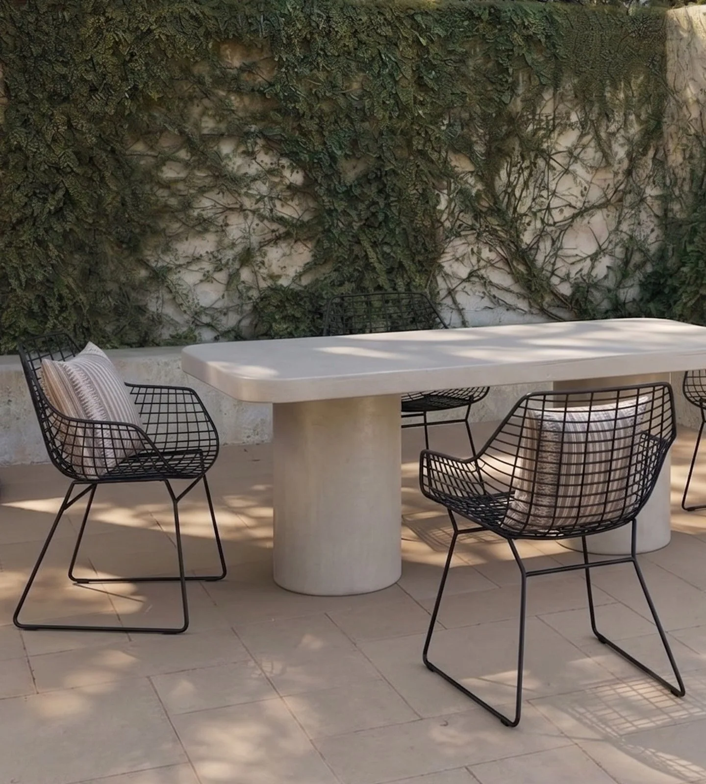 Milano rectangular white concrete dining table on twin cylindrical concrete pedestals in a sunlit Mediterranean courtyard with black wire armchairs, striped cushions and an ivy-clad plaster wall