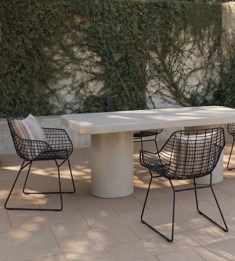 Milano rectangular white concrete dining table on twin cylindrical concrete pedestals in a sunlit Mediterranean courtyard with black wire armchairs, striped cushions and an ivy-clad plaster wall