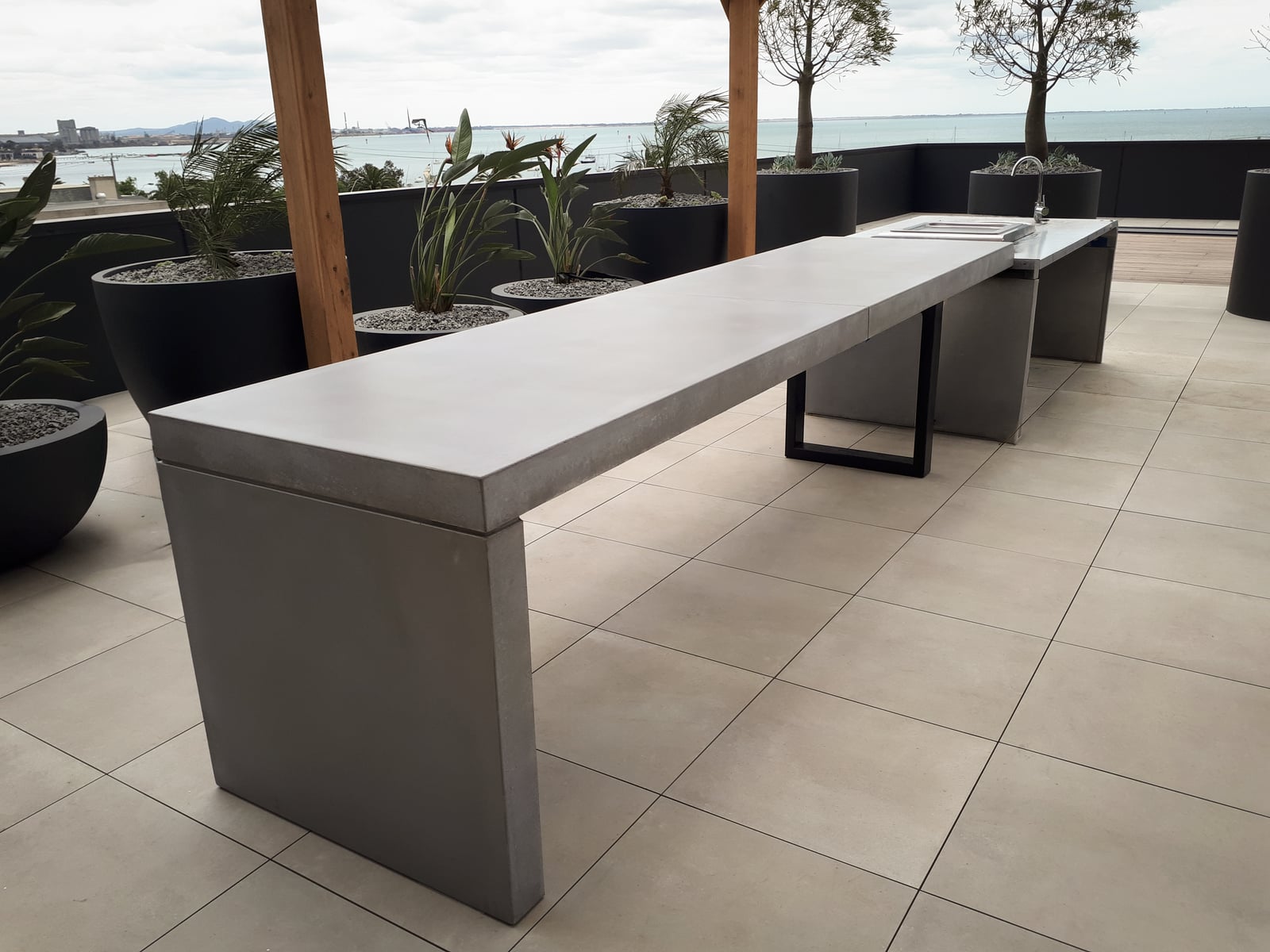 Concrete BBQ benchtop on a Melbourne rooftop terrace, paired with stainless grill station and an ocean view through palms
