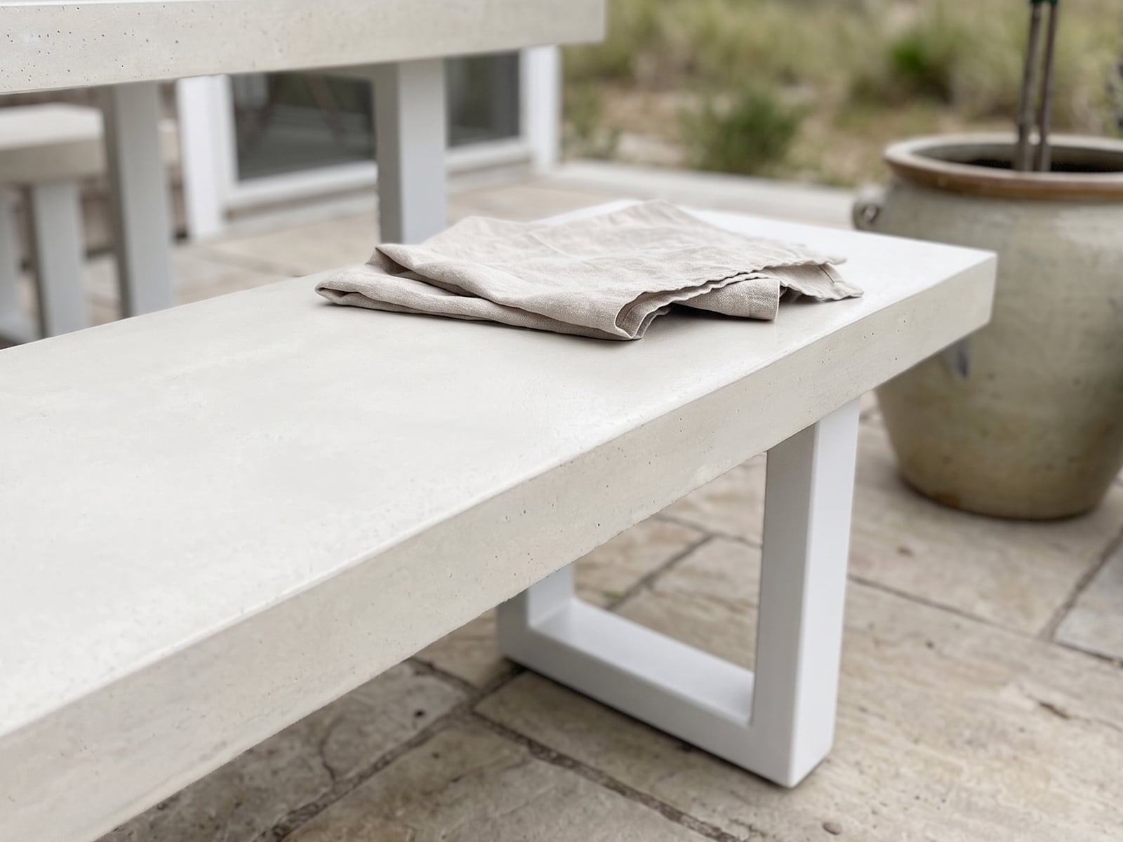 Bone white concrete outdoor bench seat on a flagstone terrace with linen napkins, framed by coastal grasses