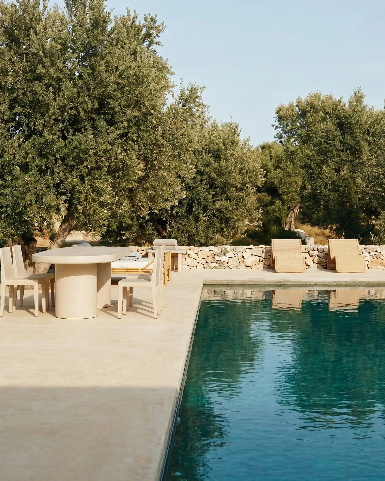 A bone white concrete oval dining table beside a Mediterranean pool, framed by olive trees and a stacked-stone wall, with rattan loungers on the far side