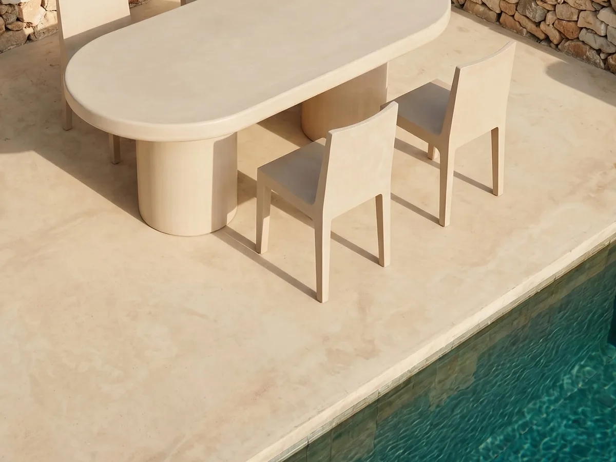 Bone-white SnapCo concrete outdoor dining table on a round concrete pedestal base beside a turquoise pool, paired with bone-coloured timber chairs