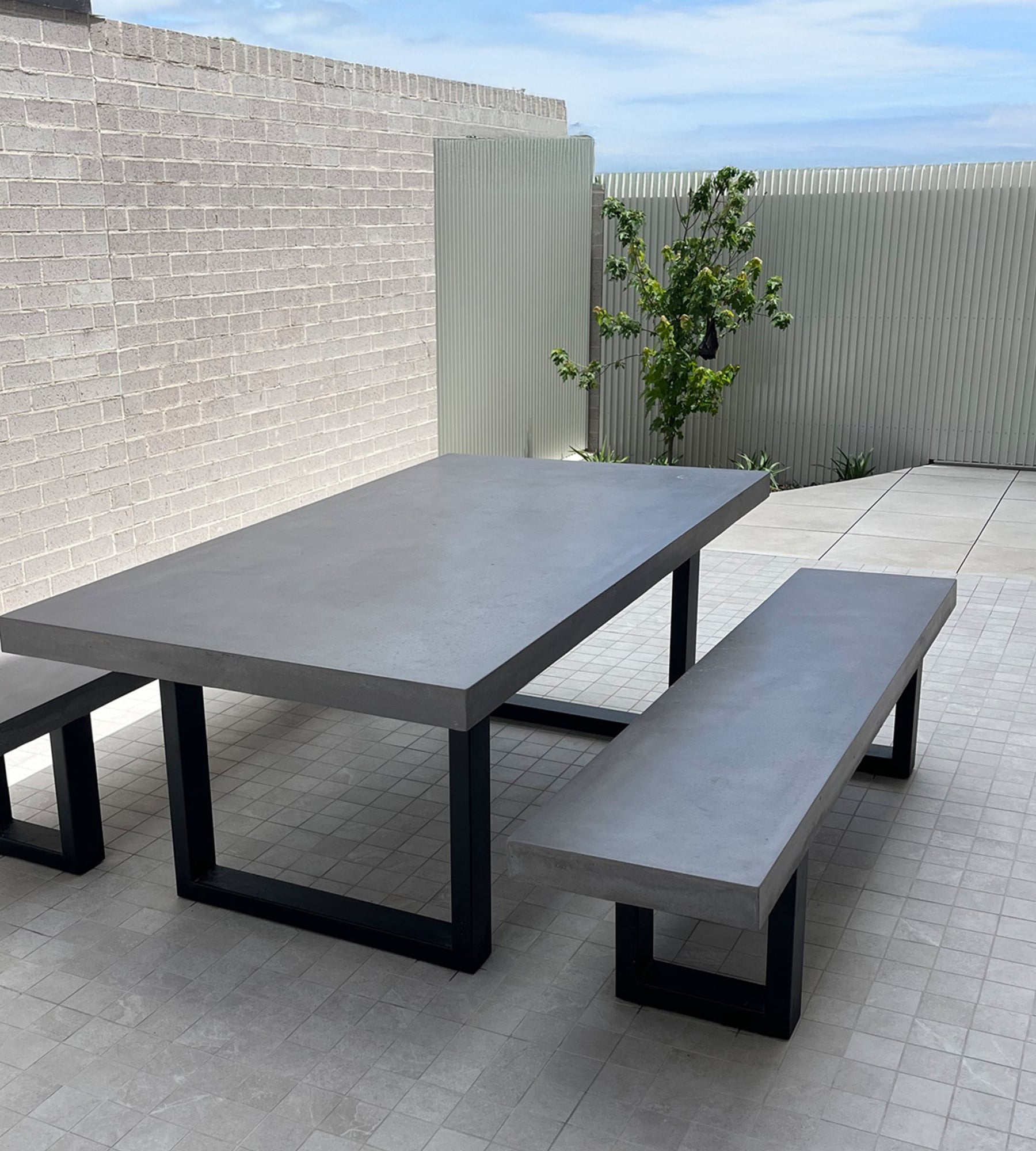 Bone-white SnapCo concrete outdoor dining table with matched bench seats on a stone-paved coastal patio, framed by cedar-shingle wall, lavender pots and ocean dunes