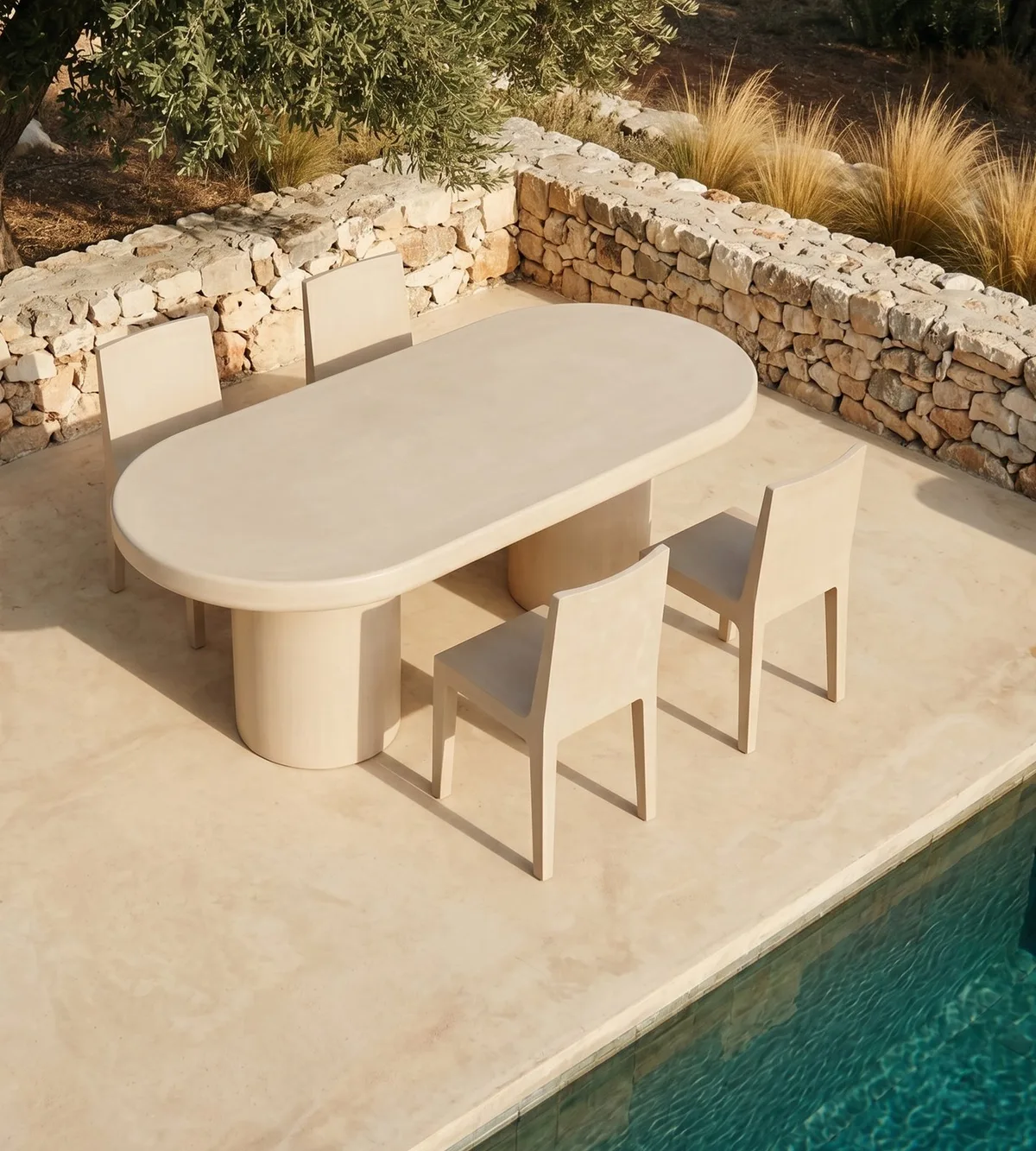 Palermo oval concrete dining table on a Mediterranean poolside terrace, parsons chairs and stacked-stone retaining wall with olive tree and golden grasses