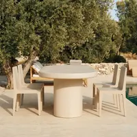 Close-up of the Palermo bone white concrete oval dining table on a poolside courtyard with sage parsons chairs, a rattan sun lounger and an old olive tree
