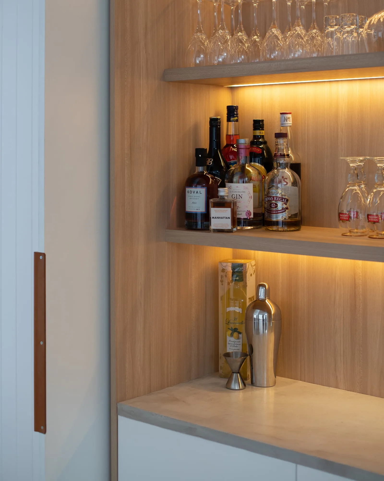 A bone white concrete bar surface set into a light timber shelving bay, glassware above and a cocktail shaker on the polished cast top
