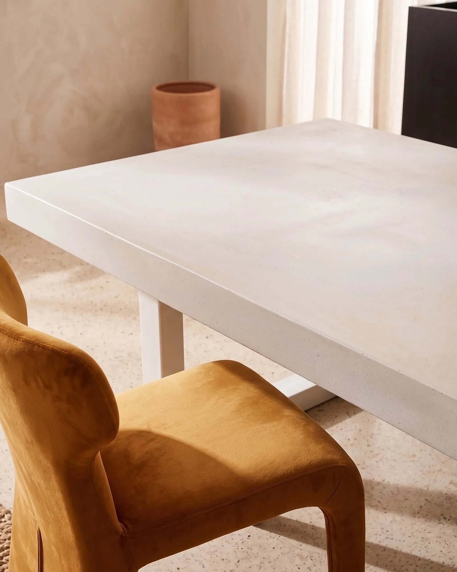 Close-up of a bone white concrete dining table with a mustard velvet chair, terracotta planter and limewash plaster wall, set on a terrazzo floor