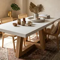 Overhead view of the TimberCross white concrete dining table with cane chairs, stoneware vessels, dried palms and a stone tray on a persian rug