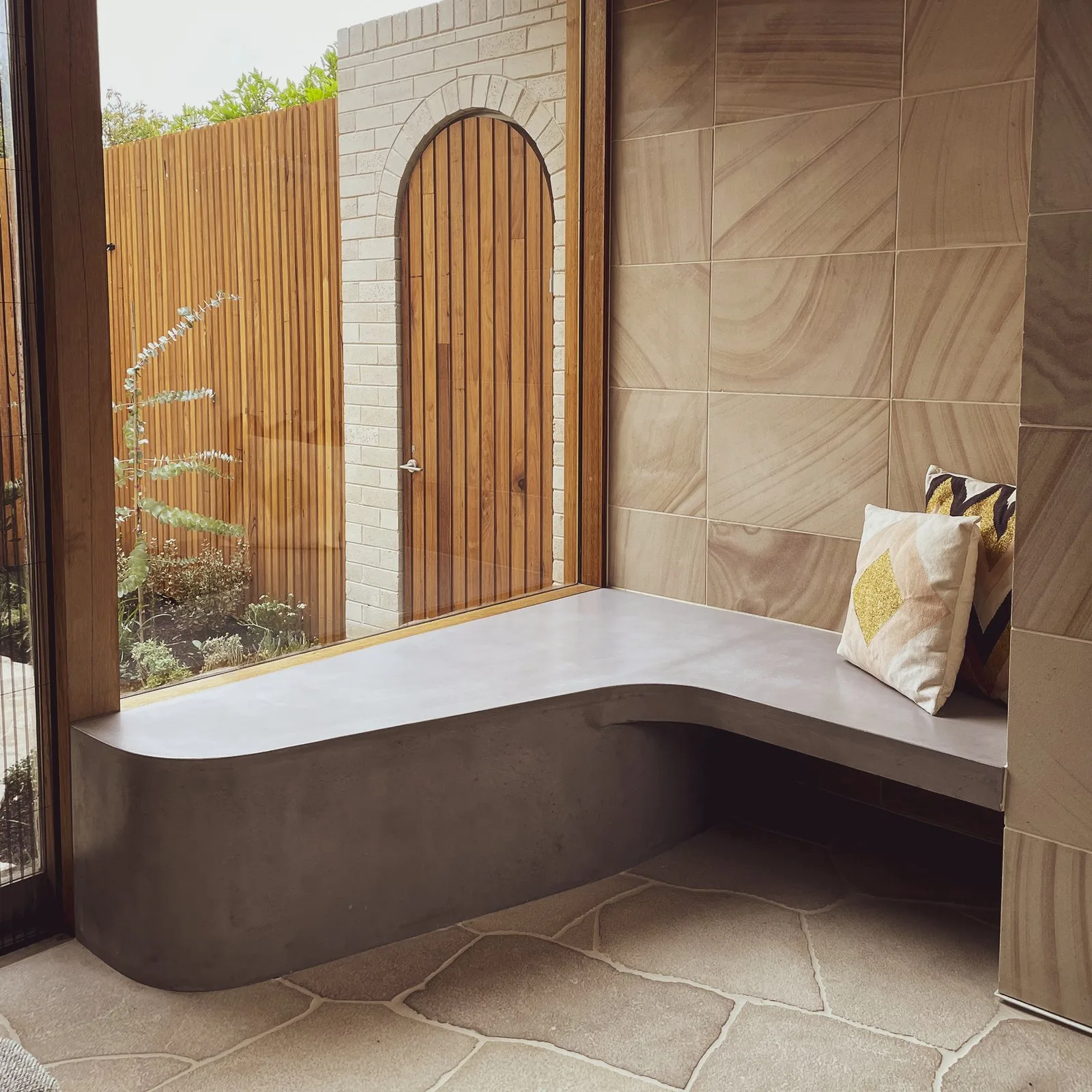 Custom-built curved concrete bench seat in a sandstone-tiled Melbourne courtyard nook, a bespoke residential commission with timber slat fencing and an arched timber door beyond
